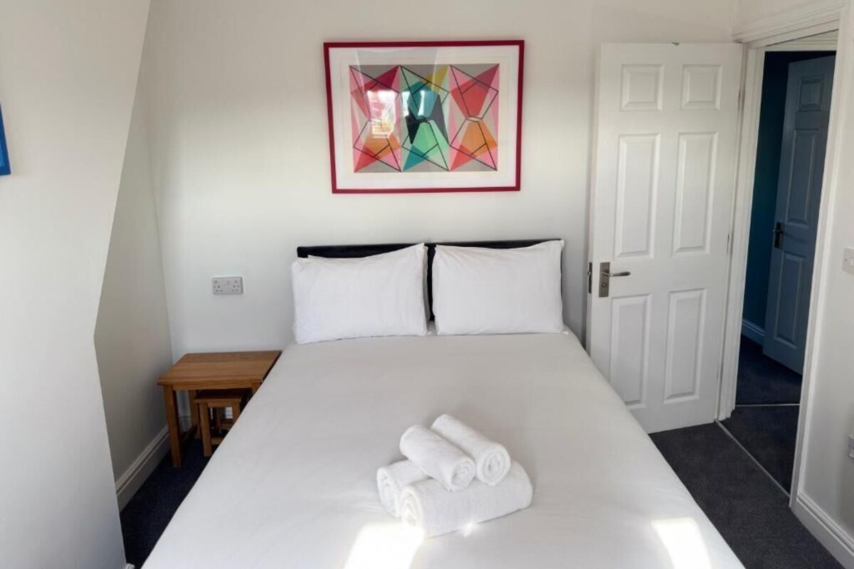 A hotel room with a bed, a small table, a framed artwork on the wall, and a door.