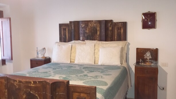 Ein Schlafzimmer mit einem Holzbettgestell, einer gemusterten Bettdecke, einem Nachttisch mit Lampe und einer an der Wand hängenden Uhr.