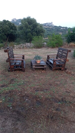 Un área de estar al aire libre con estilo rústico, con bancos de madera y una pequeña mesa, rodeada de vegetación natural y un muro de piedra.