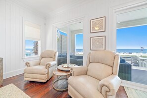 Une pièce avec deux fauteuils en cuir beige, une petite table ronde et une vue sur la plage grâce à de grandes fenêtres.