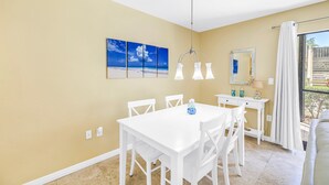 A dining area with a white table and chairs, a chandelier, and a view of the outdoors through a window. - Gertrude's Siesta | Bayview Unit, Near Beach, Private Patio, Heated Pool, Pickle,Tennis! (Siesta Key)