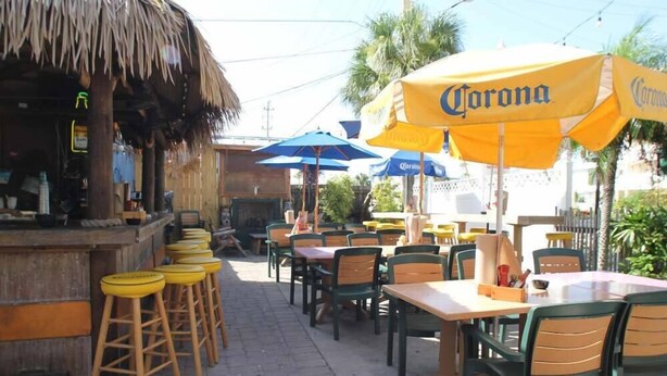 An outdoor dining area with wooden tables and chairs, yellow and blue umbrellas, and a thatched-roof structure. - Gertrude's Siesta | Bayview Unit, Near Beach, Private Patio, Heated Pool, Pickle,Tennis! (Siesta Key)