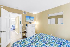 A bedroom with a bed, dresser, and a wall-mounted TV displaying a beach scene. - Gertrude's Siesta | Bayview Unit, Near Beach, Private Patio, Heated Pool, Pickle,Tennis! (Siesta Key)