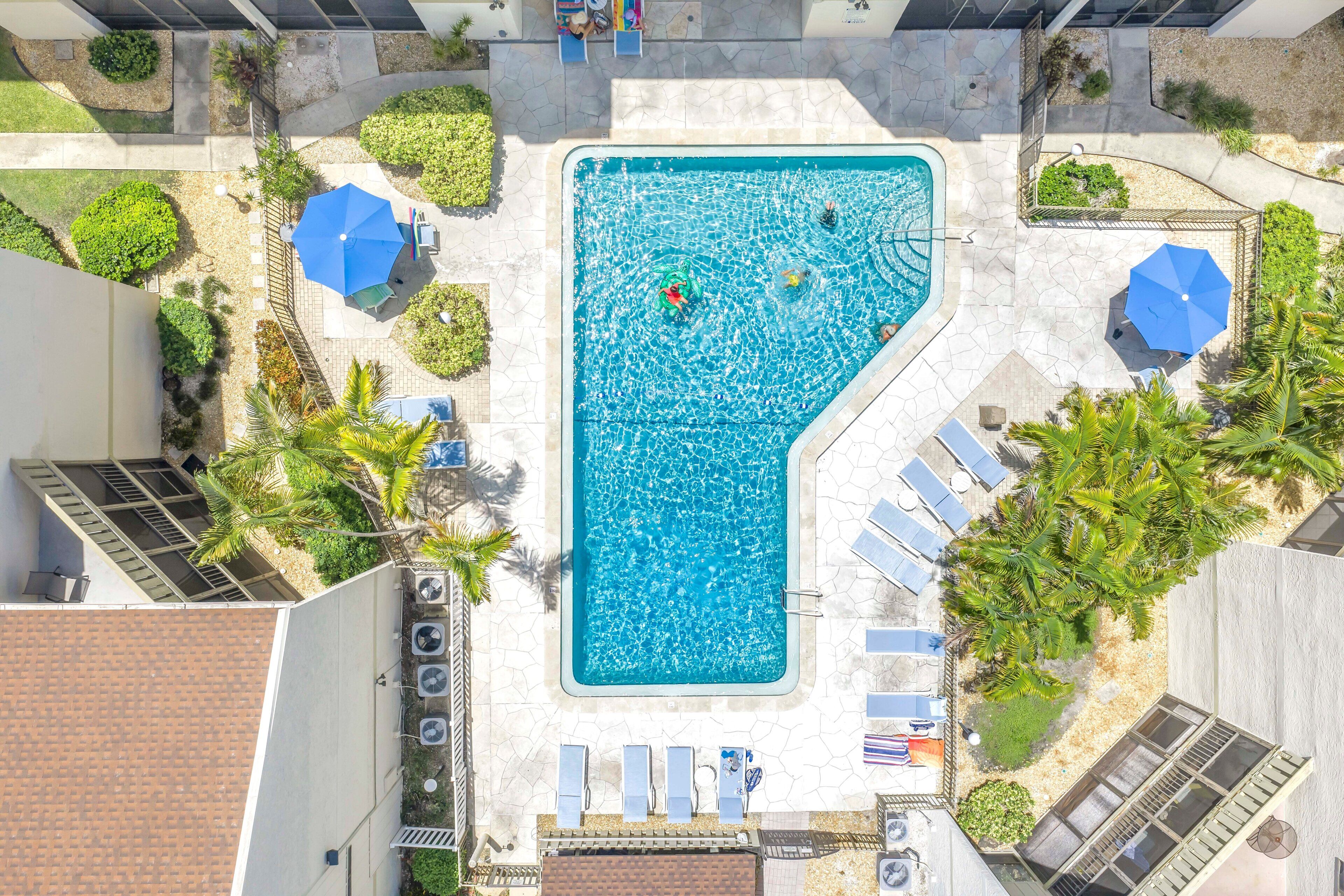 Vista aérea de una piscina rodeada de sillas de descanso y sombrillas, bajo un cielo despejado.