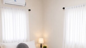 Et soveværelse med seng, hvide gardiner, et natbord med lampe og en aircondition.