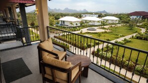 A balcony with a wooden chair and table, overlooking a landscaped garden with white tents and mountains in the distance.