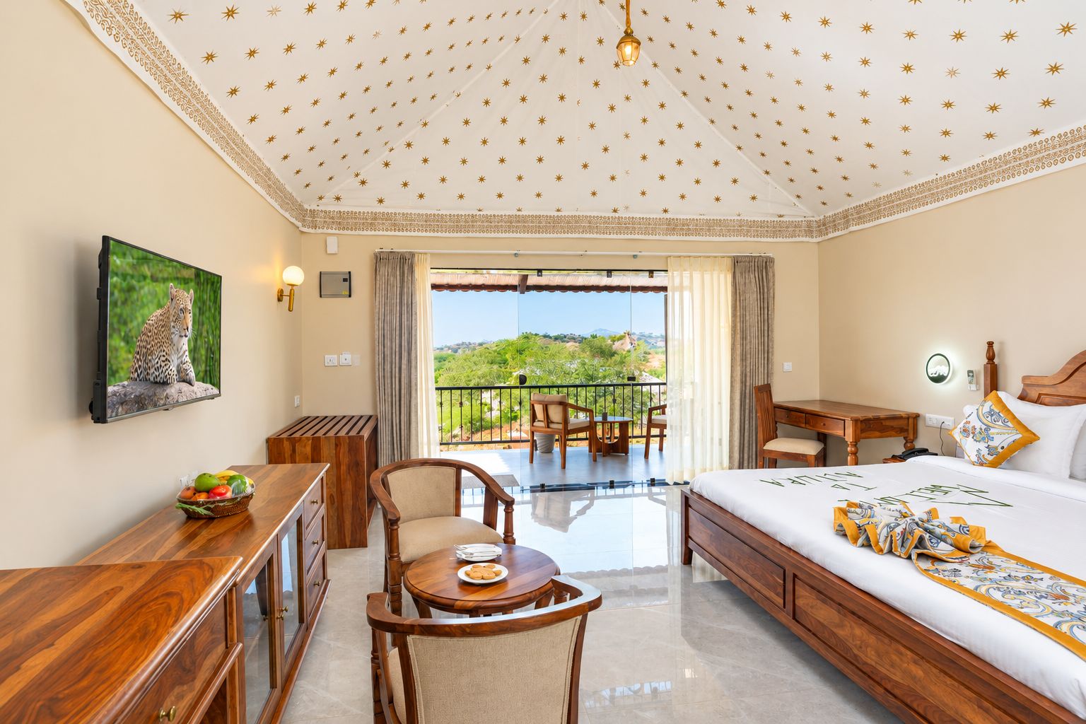 A hotel room with a balcony, a bed with a patterned bedspread, a desk with a chair, a television, and a fruit basket.
