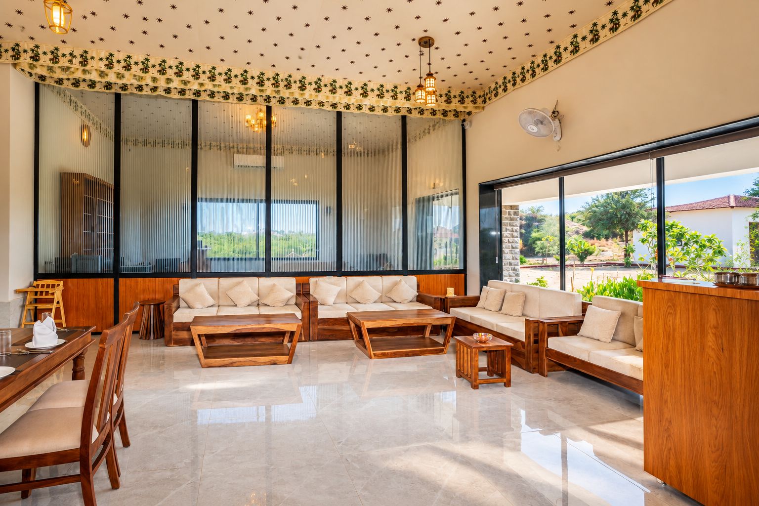 A spacious living area with a sofa set, wooden coffee tables, and a dining table with chairs. Large windows provide a view of the outdoors.