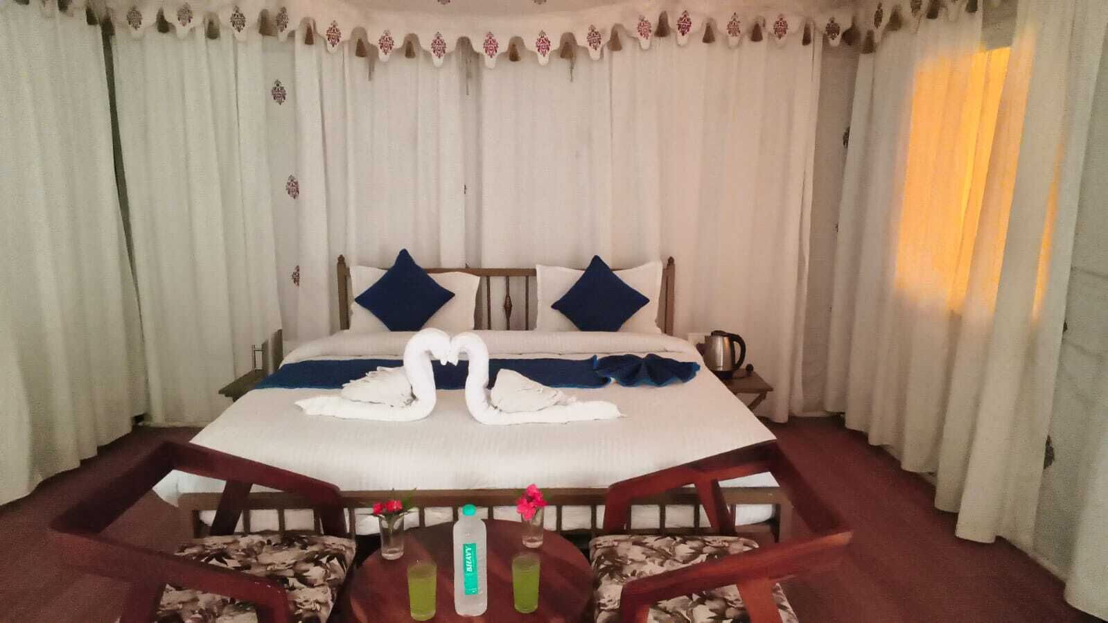 A hotel room with a bed, two pillows, a bedside table, a chair, and a table with two bottles.