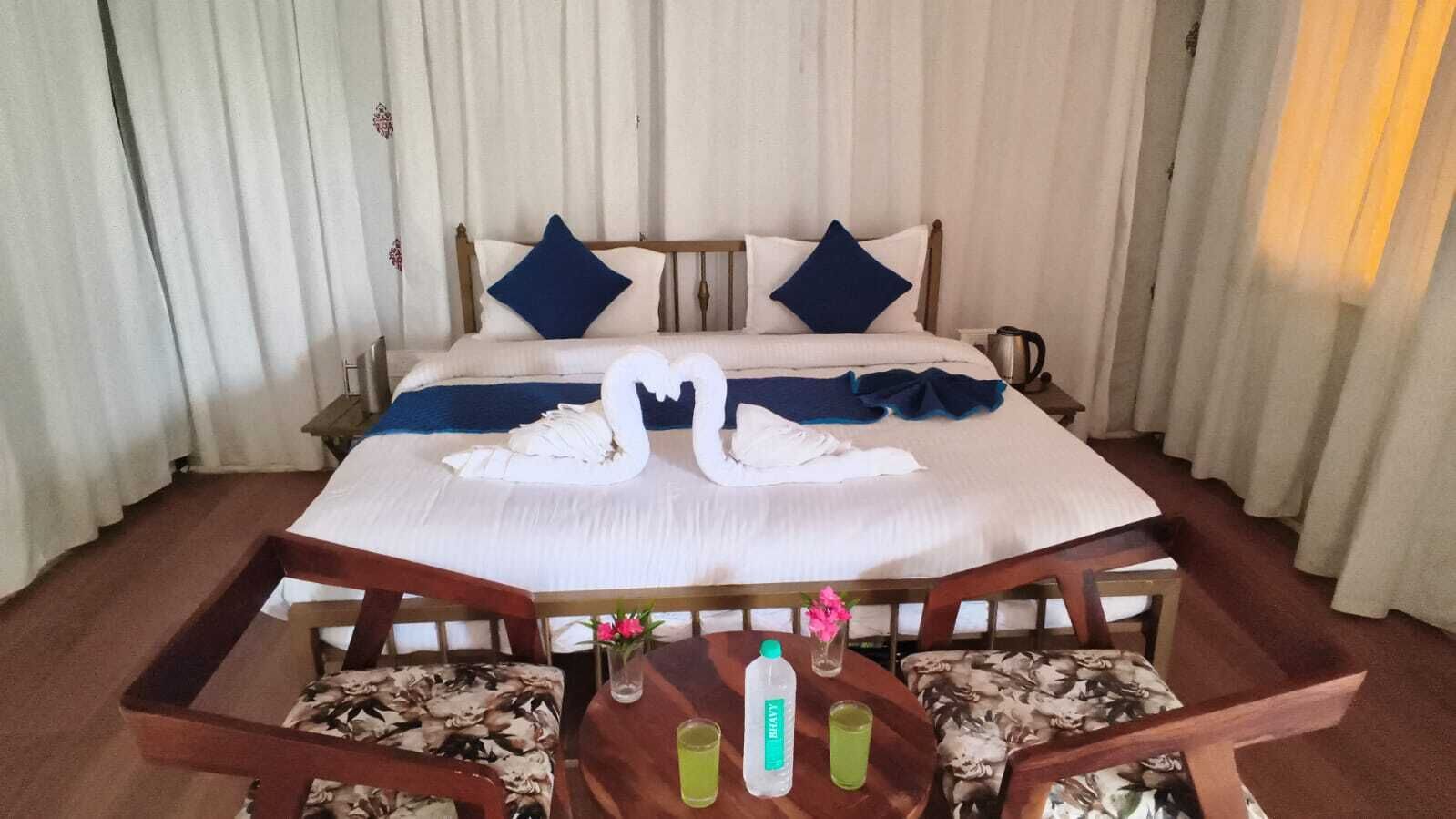 A hotel room with a bed, two pillows, a swan-shaped towel arrangement, a bedside table with a bottle and a glass, and a chair with a floral pattern.