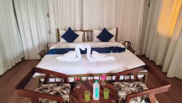 A hotel room with a bed, two pillows, a swan-shaped towel arrangement, a bedside table with a bottle and a glass, and a chair with a floral pattern.