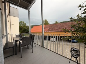 Ein Balkon mit Tisch und Stühlen, ein Metallzaun und ein Gebäude mit rotem Dach im Hintergrund.
