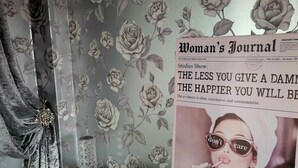 Una habitación con papel tapiz floral, un periódico sobre un atril y un jarrón decorativo.