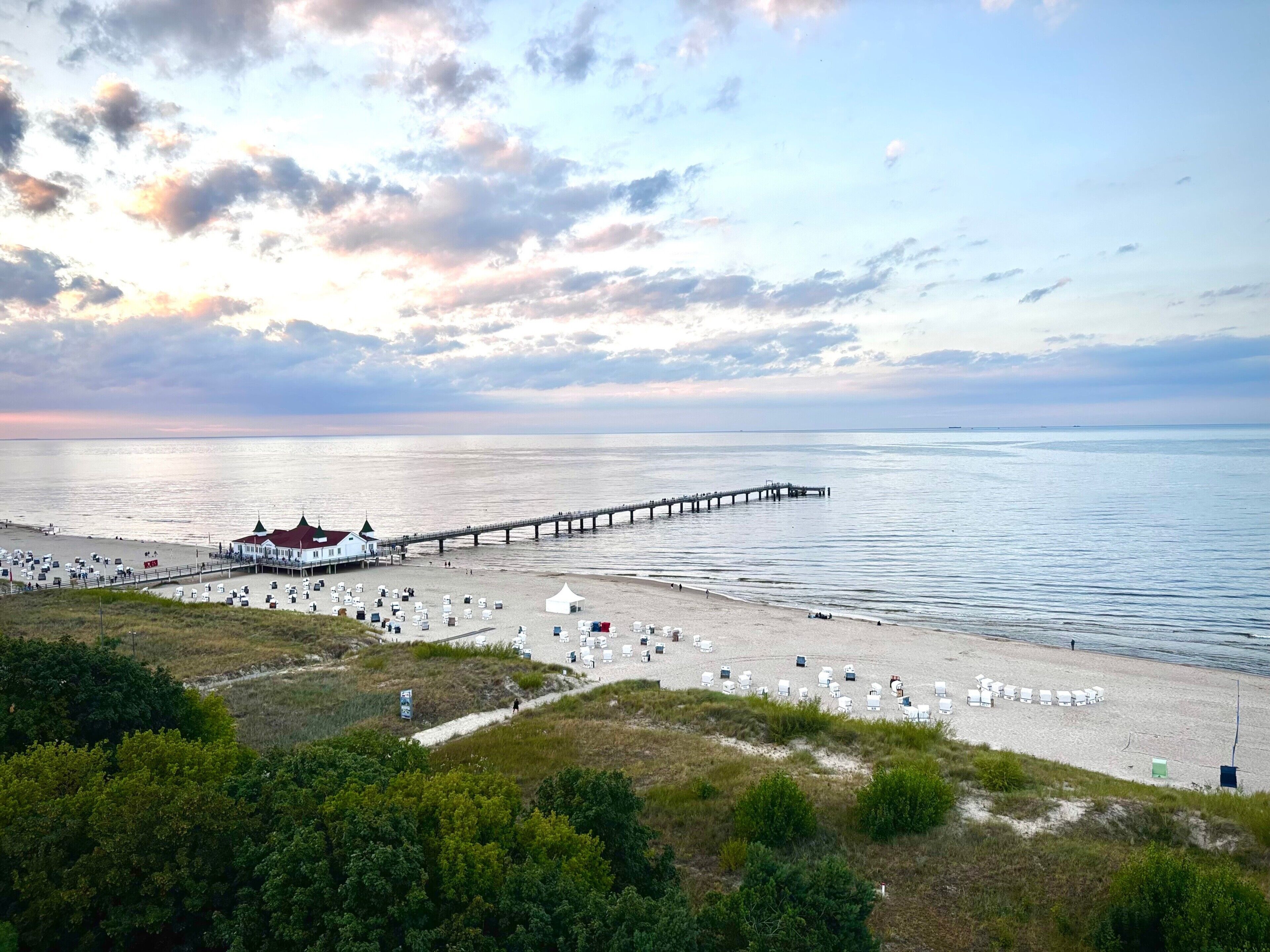 A beach with a pier extending into the water, a white building with a green roof, and numerous beach chairs and umbrellas.