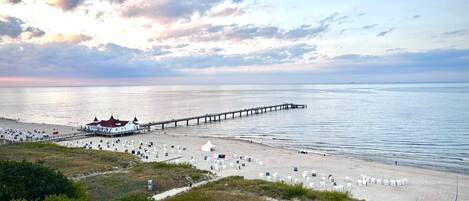 A beach with a pier extending into the water, a white building with a green roof, and numerous beach chairs and umbrellas.