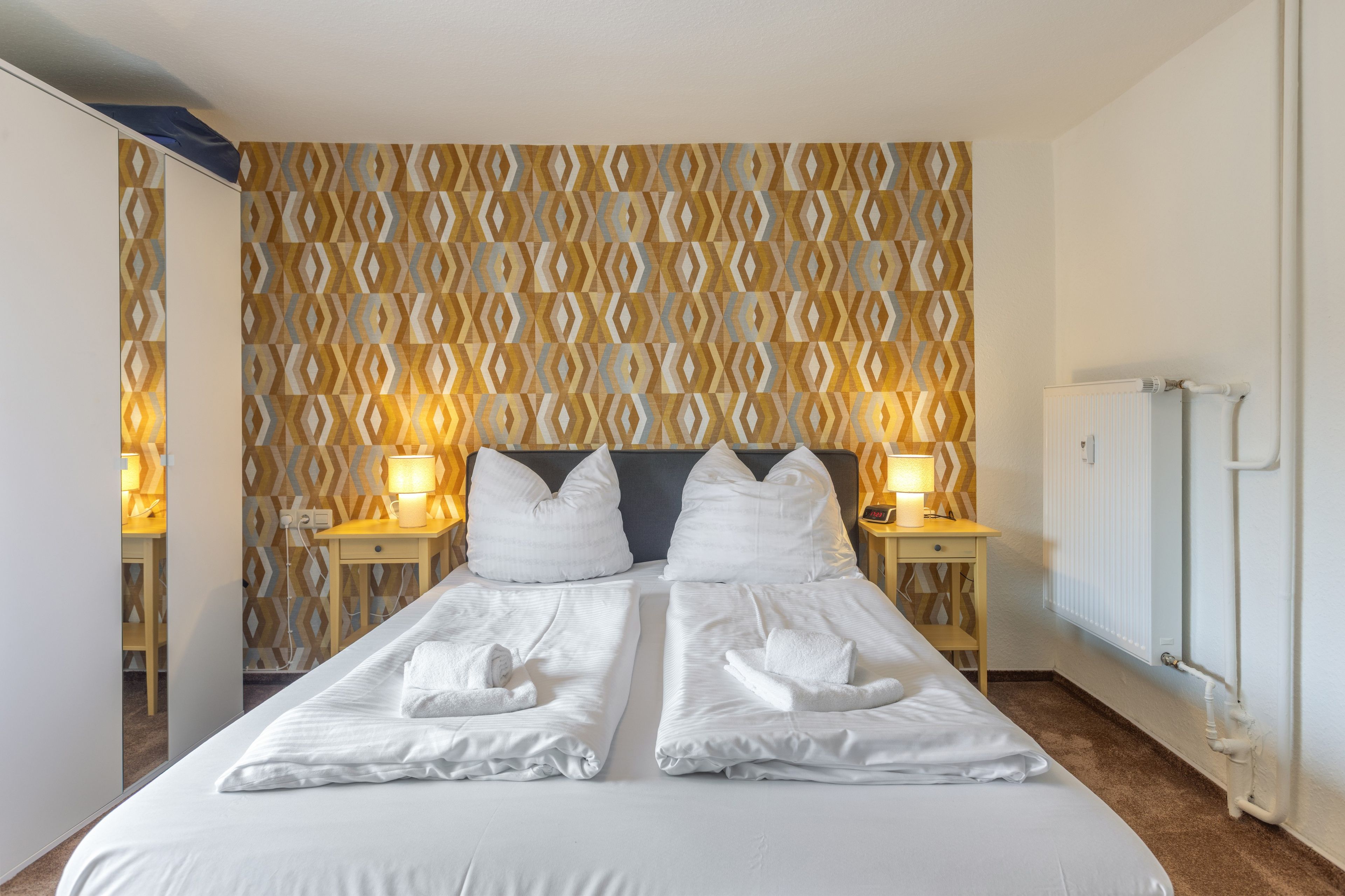 Una habitación de hotel con una cama, dos lámparas de noche, una pared con un diseño geométrico y un radiador.