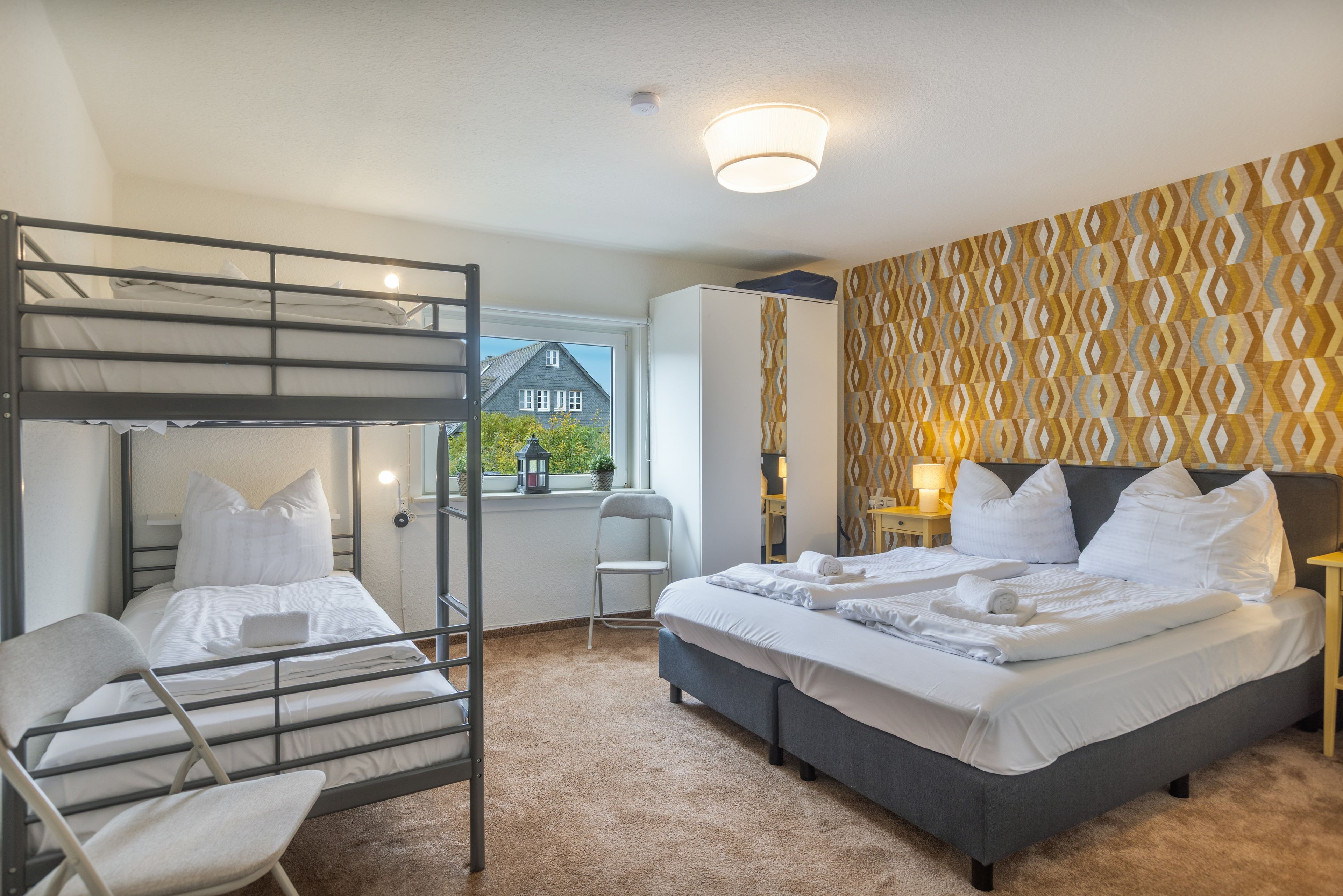 Une chambre avec des lits superposés, un lit simple, une chaise, une fenêtre donnant sur une maison et un papier peint à motifs.