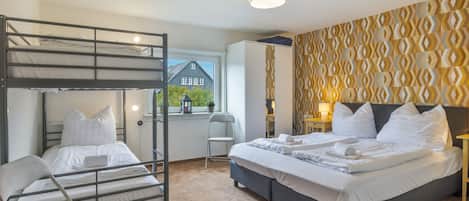 A room with bunk beds, a single bed, a chair, a window with a view of a house, and patterned wallpaper.