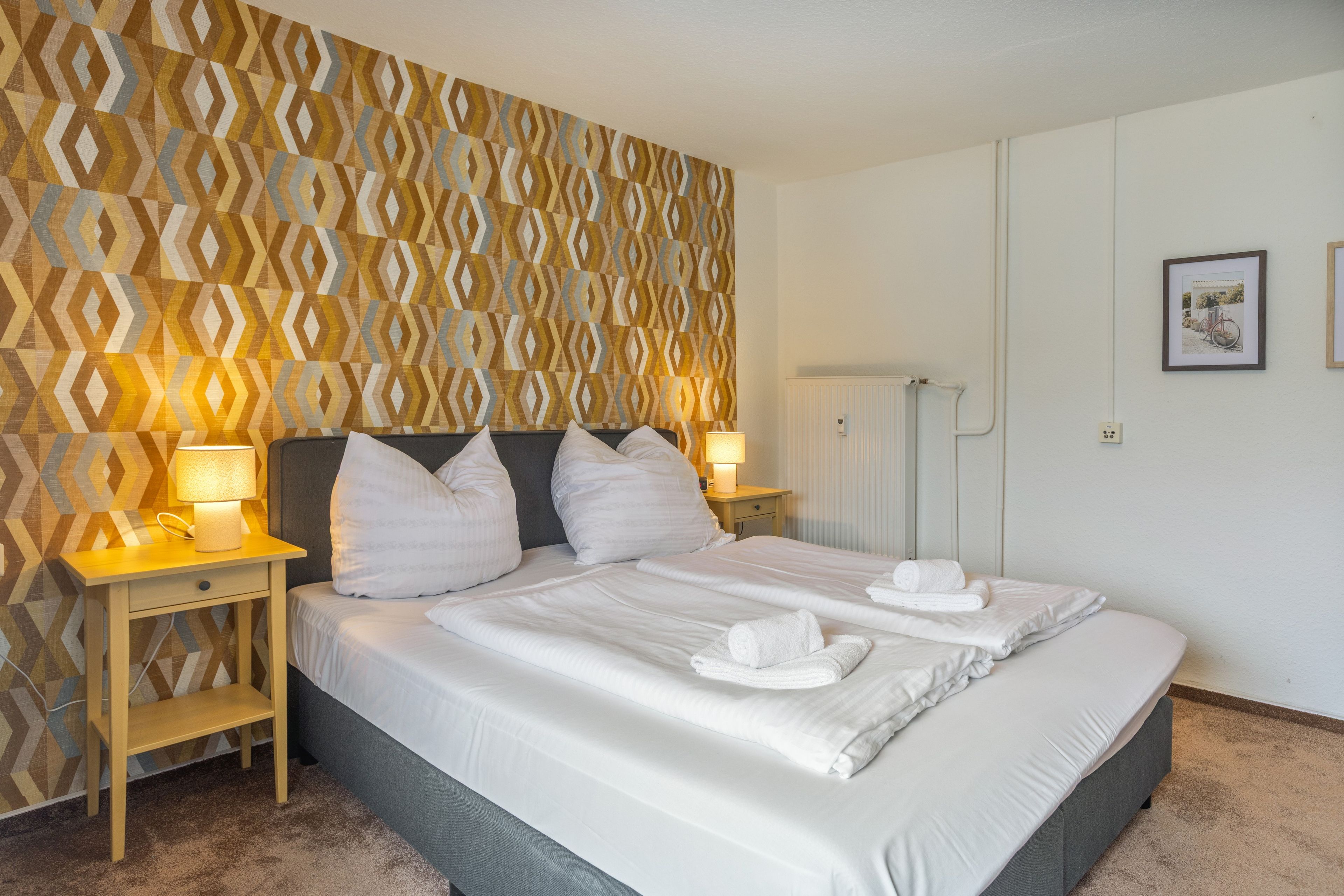 A hotel room with a patterned wallpaper, a bed with white linens, two bedside lamps, and a framed picture on the wall.