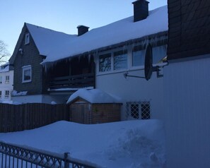 Ein Haus mit geneigtem, mit Schnee bedeckten Dach, an dem Eiszapfen von den Dachvorsprüngen hängen, und einer Satellitenschüssel an der Seite.