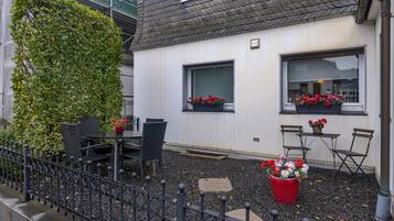 Un edificio con fachada blanca, dos ventanas con macetas de flores, un pequeño espacio al aire libre con mesa y sillas, y una cerca de metal.