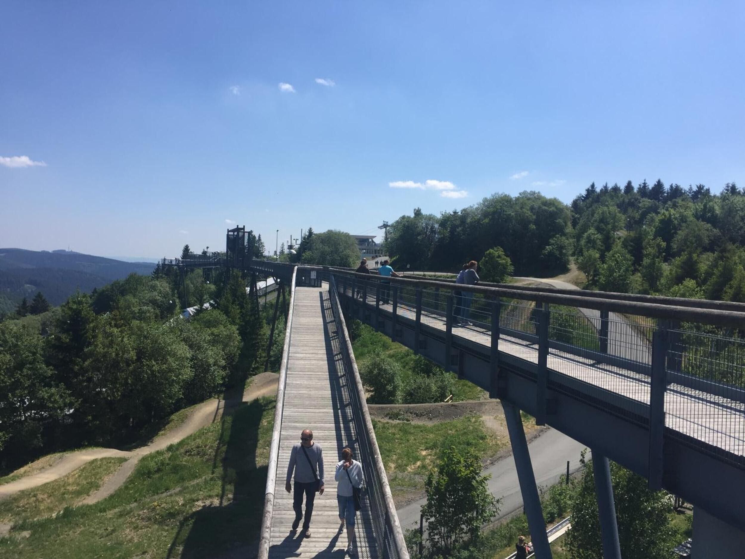 Un puente peatonal con barandilla metálica que conecta dos zonas elevadas, con una carretera debajo.