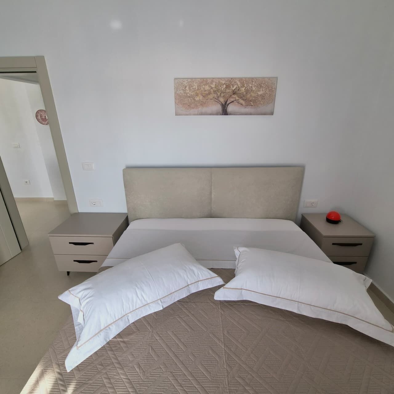 Un dormitorio con una cama, dos mesitas de noche, una mesita auxiliar, una obra de arte en la pared y una puerta.
