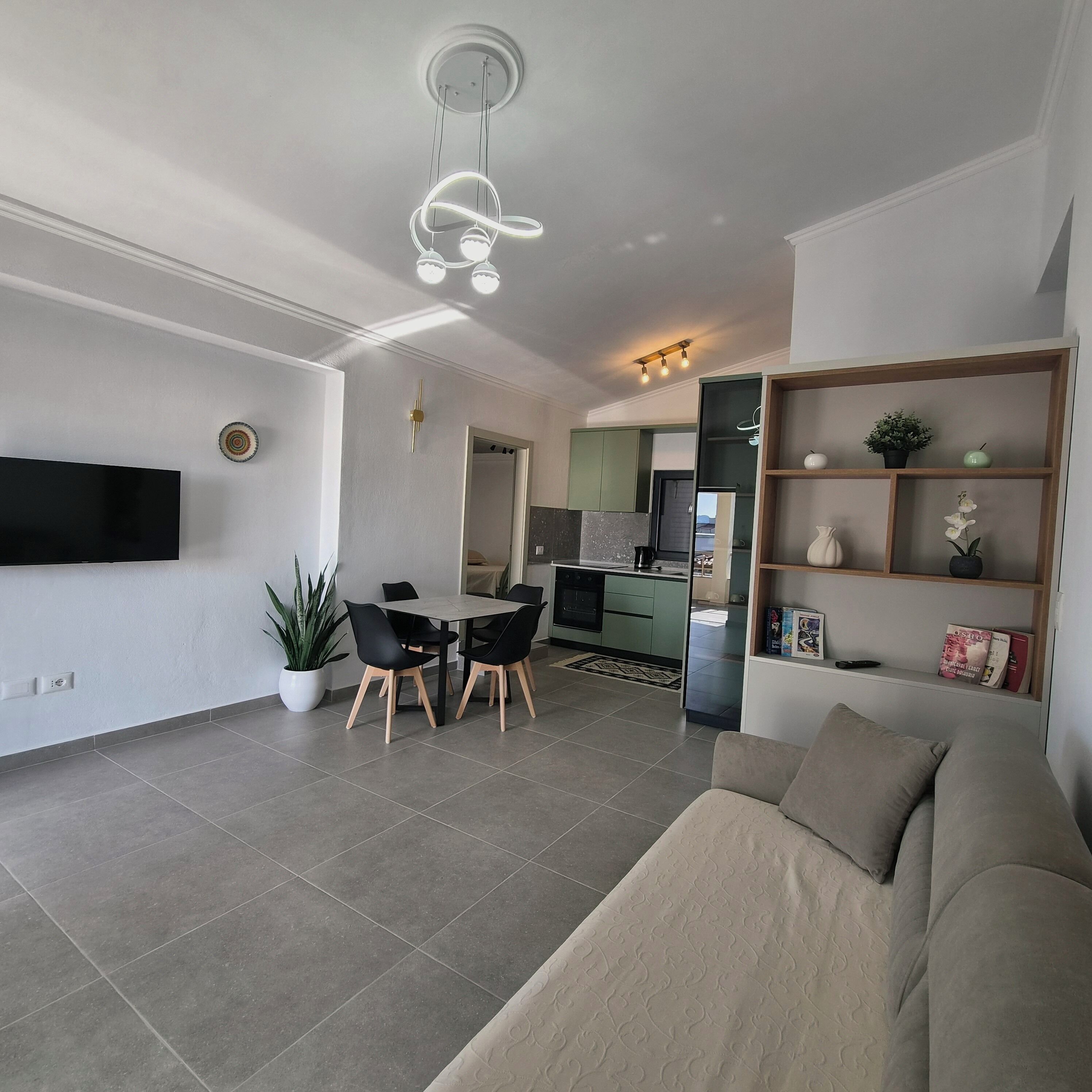 A modern living room with a sofa, dining table, chairs, a flat-screen TV, and a kitchen in the background.