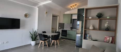 A modern living room with a sofa, dining table, chairs, a flat-screen TV, and a kitchen in the background.