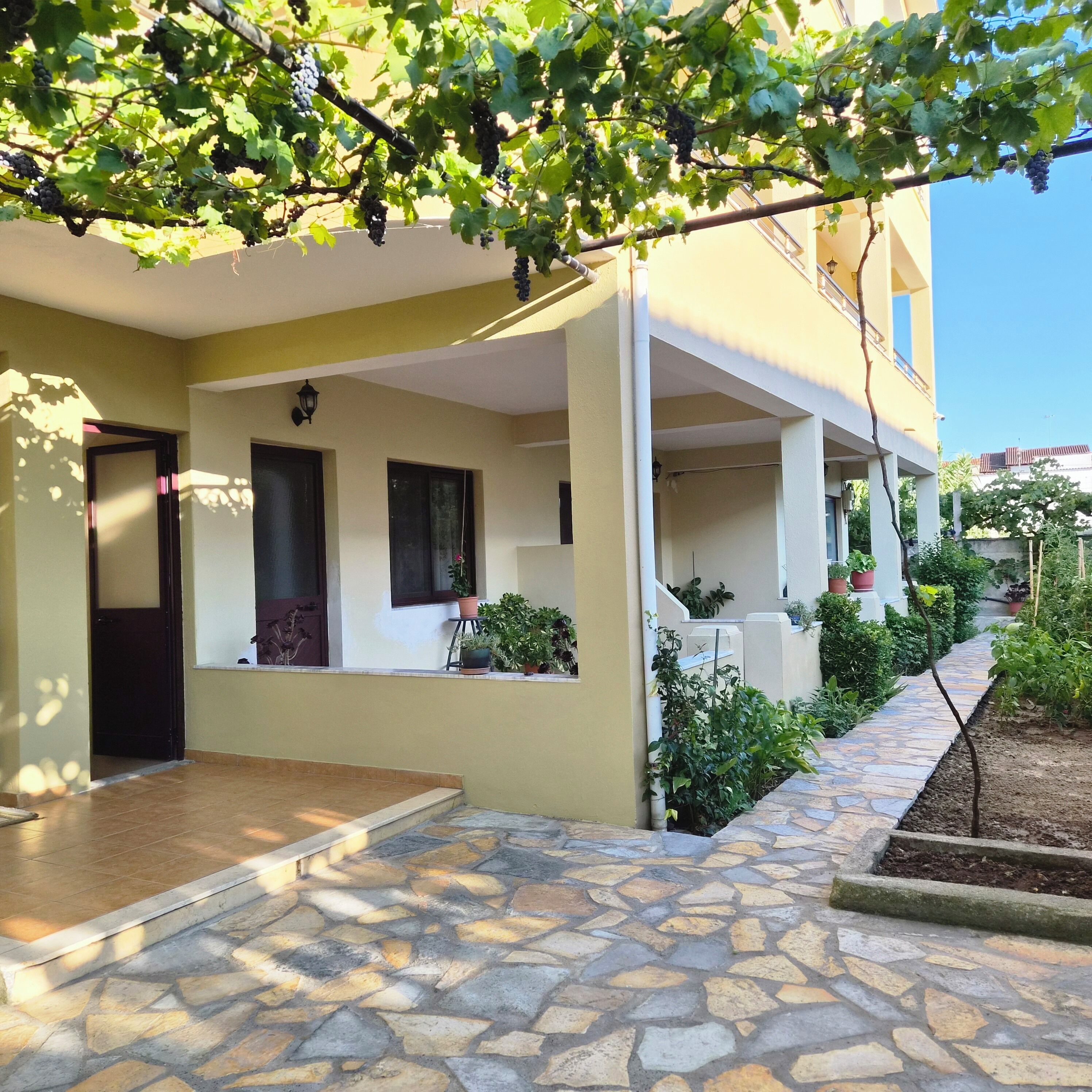 A covered outdoor area with a stone floor, potted plants, and a building with a yellow facade.