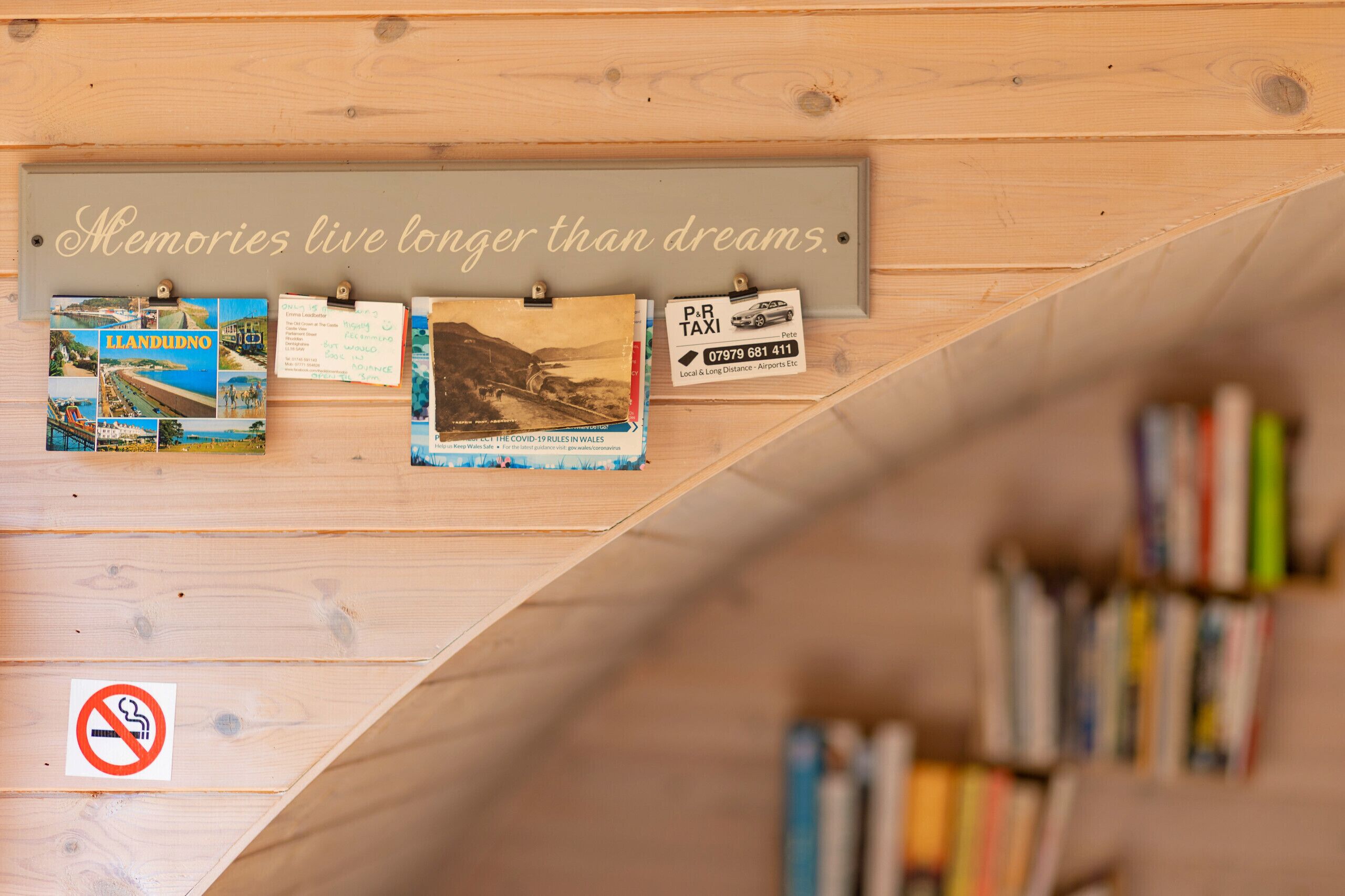 A wooden wall with a plaque that reads 'Memories live longer than dreams.' Below, there are several postcards pinned to the wall, and a no-smoking sign is visible.