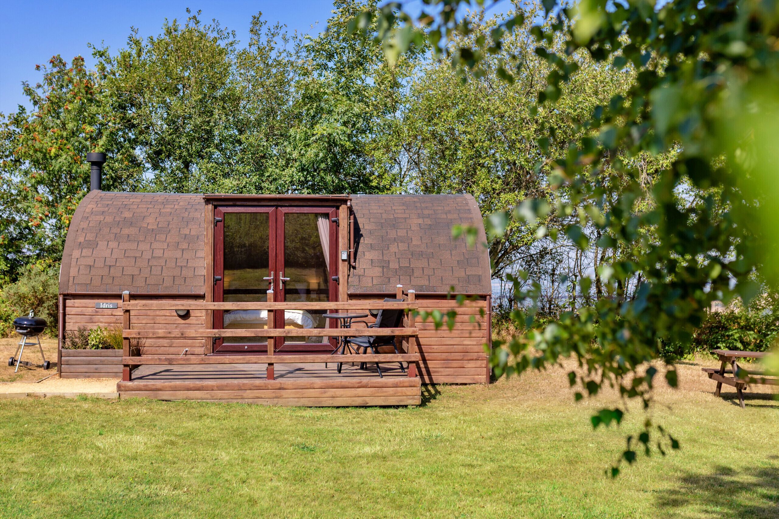 Una cabaña de madera con techo de ladrillo, puerta de cristal y una pequeña terraza con asientos.