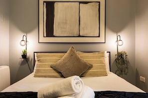 A neatly made bed with a striped pillow and a rolled towel, a framed abstract painting above, and two wall-mounted lamps on either side. - Pelican House One | Luxury Apartment | 6 Min Walk to Beach | Bournemouth Centre (Bournemouth)