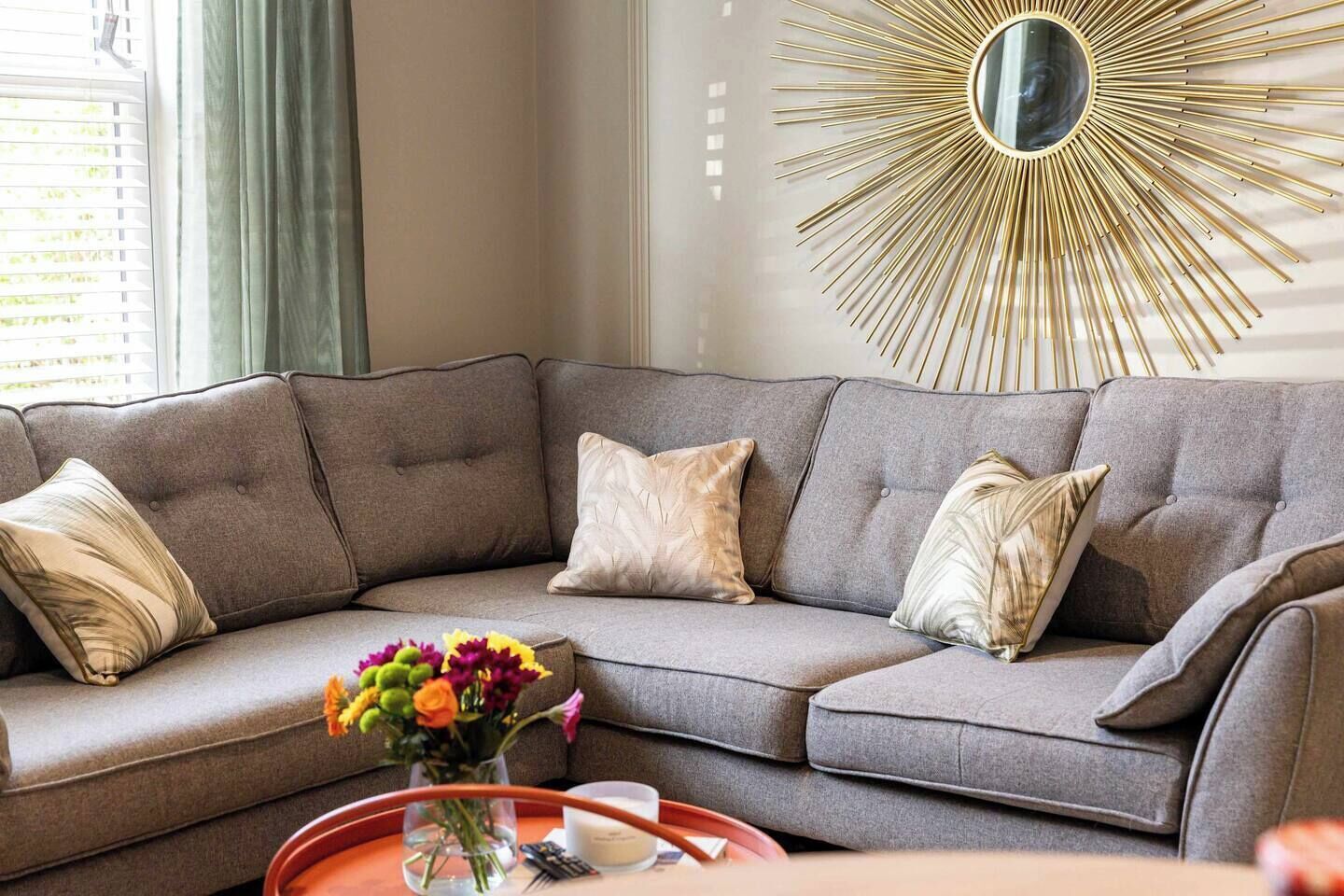 A modern living room with a sectional sofa, a decorative sunburst mirror, and a vase with flowers on a side table.