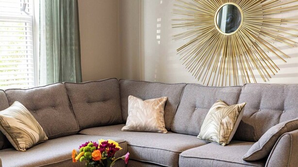 Un soggiorno moderno con un divano componibile, uno specchio decorativo a forma di sole e un vaso con fiori su un tavolino.