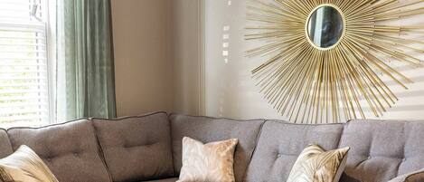 A modern living room with a sectional sofa, a decorative sunburst mirror, and a vase with flowers on a side table.