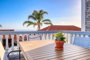 Ein Balkon mit einem Holztisch, einem Blumentopf und Blick auf eine Palme sowie rote Ziegeldächer.