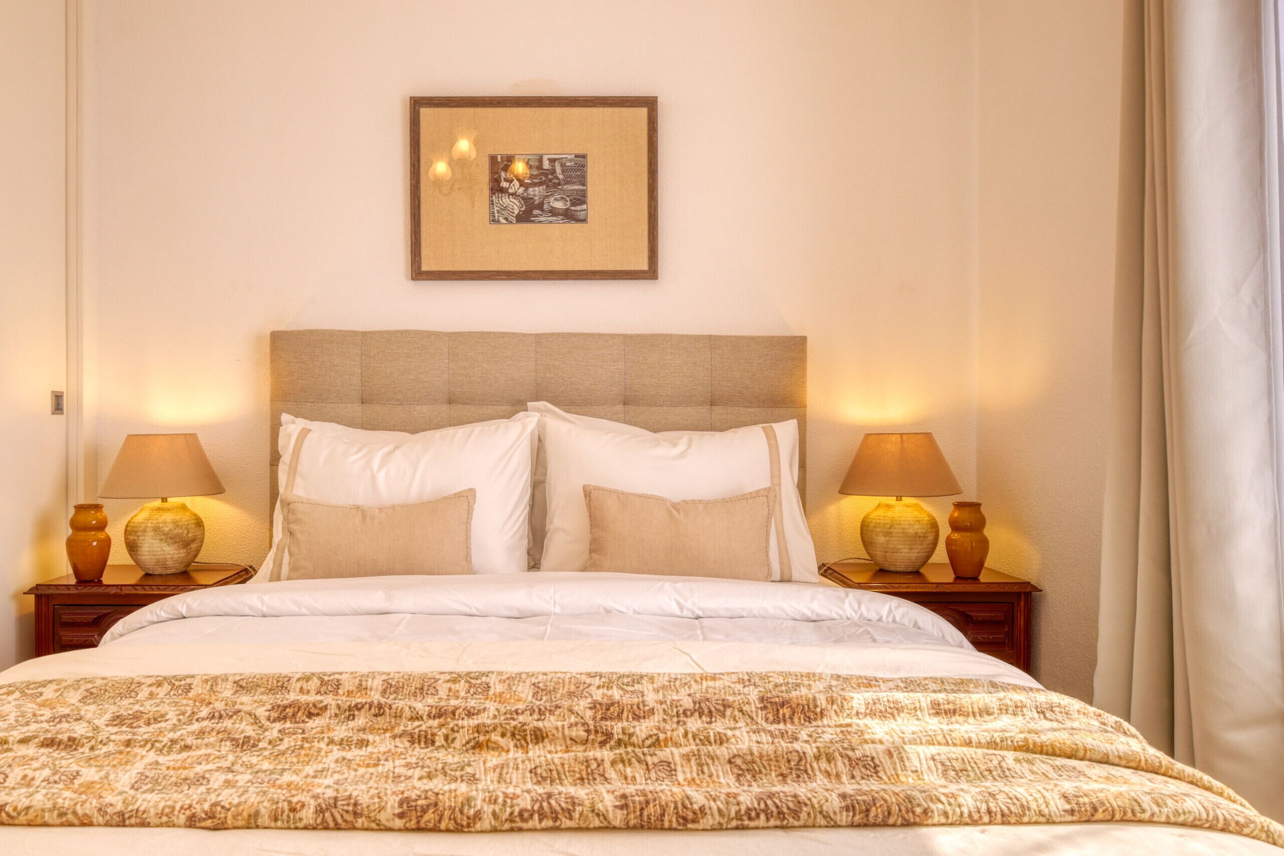 Une chambre d’hôtel avec un lit, deux lampes de chevet, un tableau encadré au mur et une fenêtre avec des rideaux.