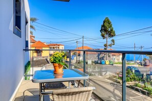 A balcony with a table and chair, a potted plant, and a view of the street with buildings and parked cars. - Casa do Serrado, a Home in Madeira (Ponta do Sol)