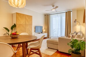 A modern living room with a dining area, a flat-screen TV, a ceiling fan, and a large window with curtains. - Cozy Santa Cruz, a Home in Madeira (Santa Cruz)