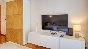 A modern living room with a flat-screen TV mounted on a white cabinet, a wooden wardrobe, and a small table with a lamp and remote controls. - Cozy Santa Cruz, a Home in Madeira (Santa Cruz)