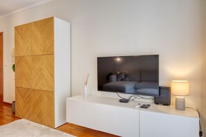 Ein modernes Wohnzimmer mit einem flachbildfernseher an einem weißen Schrank, einem Holzschrank und einem kleinen Tisch mit Lampe und Fernbedienungen.