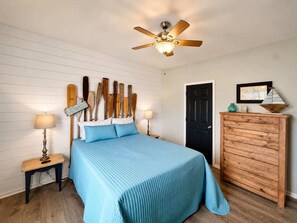 Une chambre à coucher comprenant un lit, une tête de lit en bois, un ventilateur de plafond, une table de chevet, une lampe, une commode et une porte.