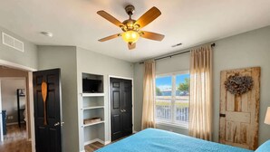 Une chambre à coucher avec un ventilateur de plafond, un lit recouvert d’une couverture bleue, une armoire et une vue par la fenêtre.