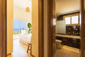 Ein Schlafzimmer mit Bett, einem kleinen Hocker, einer Pendelleuchte und Blick aufs Meer.