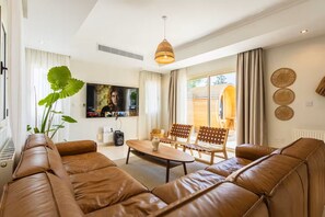 Een moderne woonkamer met een leren bank, houten stoelen, een salontafel en een tv die aan de muur is bevestigd.