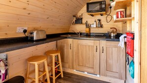 Une cuisine compacte avec des meubles en bois, un grille-pain en acier inoxydable, une bouilloire et un four à micro-ondes. On y trouve deux tabourets en bois et une couverture anti-feu accrochée au mur.