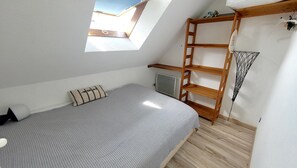 Ein kleines Zimmer mit einem Einzelbett, geneigter Decke, einem Oberlicht, einer Holzleiter und einer Heizung.