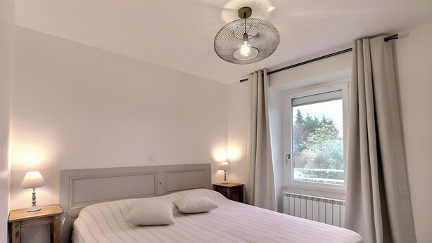 A bedroom with a bed, bedside lamps, a ceiling light fixture, a window with curtains, and a view of trees outside. - Cottage 'Gîte Le Lac Du Vert' with Lake View, Wi-Fi and Air Conditioning (Cheminas)