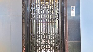 Um portão de metal com padrão de losangos, um painel de controle de elevador fixado na parede e uma parede revestida de azulejos.
